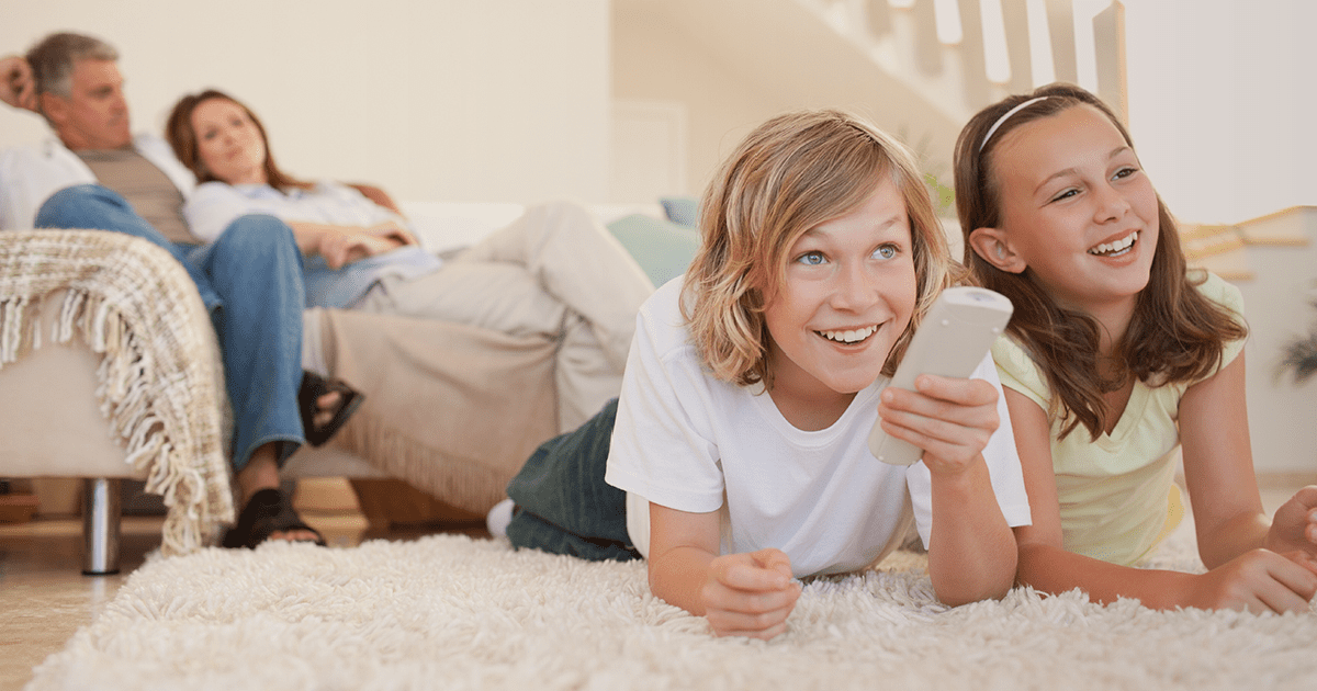 Two kids on the floor watching TV with parents in the background.