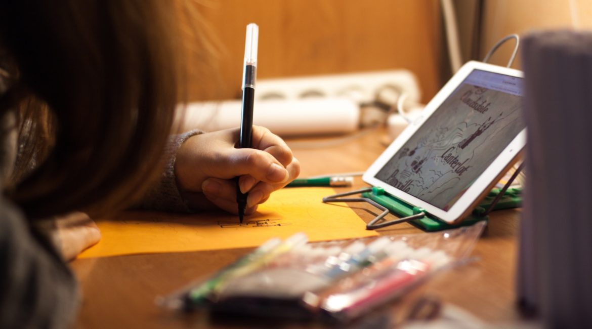 Child drawing on paper with iPad on desk.