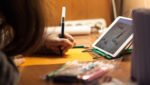 Child drawing on paper with iPad on desk.