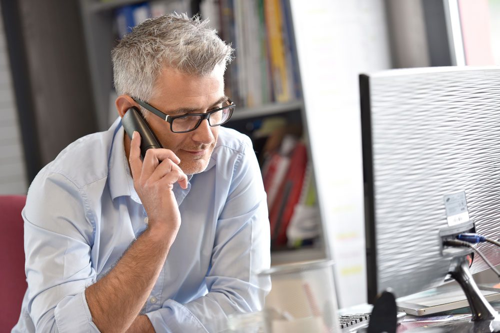 Man using landline telephone sits at his desk in front of computer monitor.