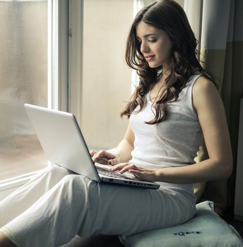 Woman in white clothes sits next to window using laptop computer.