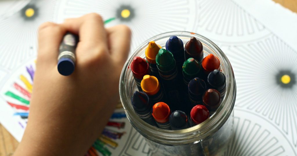 Child’s hand coloring next to jar of crayons.