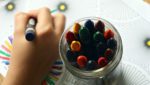 Child’s hand coloring next to jar of crayons.