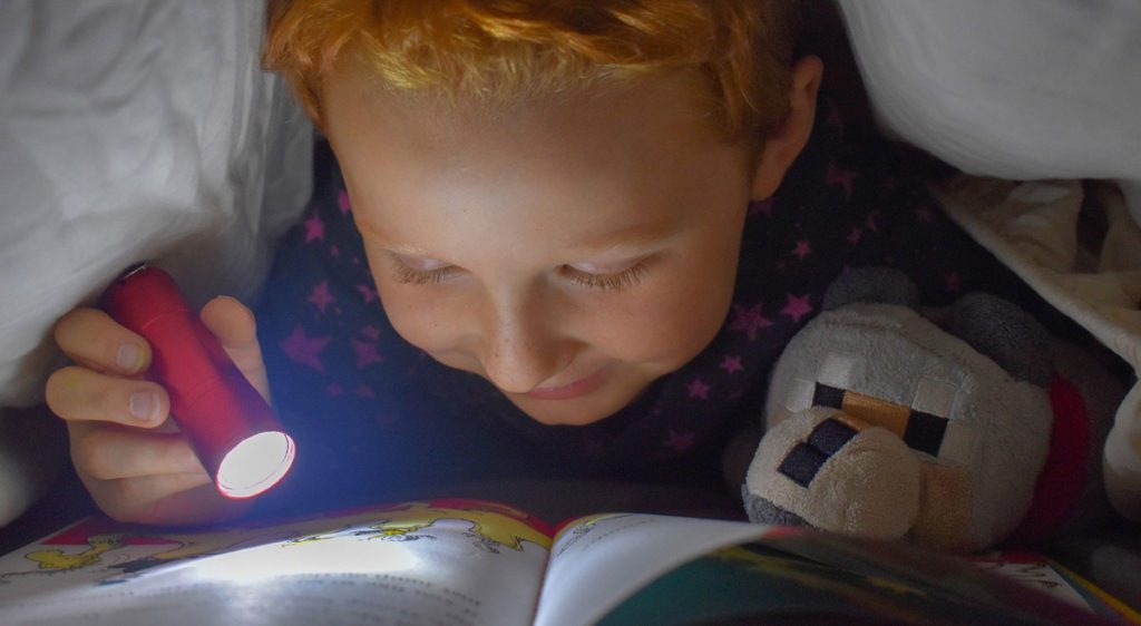 Small boy reading book in bed with flashlight.