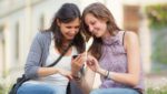 Two young women smiling and looking at a mobile phone together outside