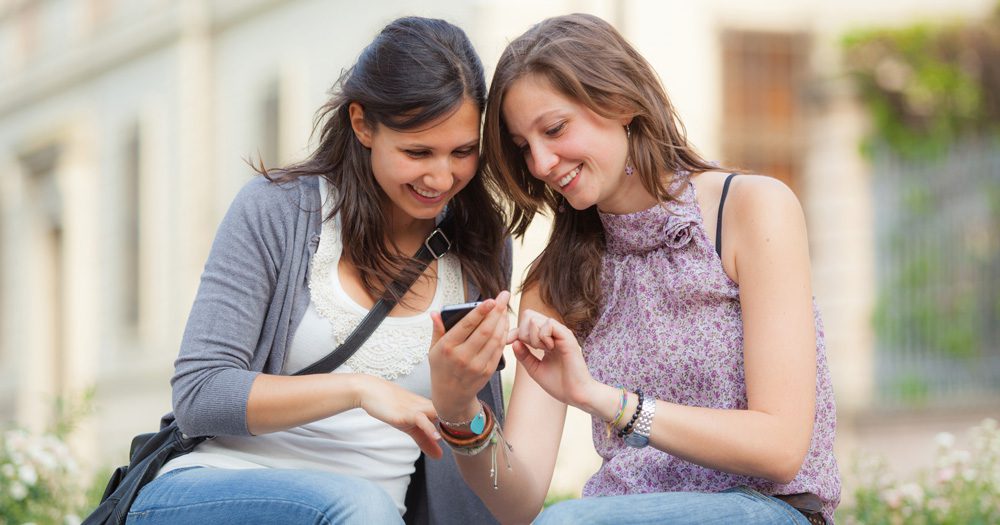 Two young women smiling and looking at a mobile phone together outside