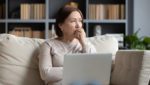 Woman on a sofa holding a laptop, looking concerned.