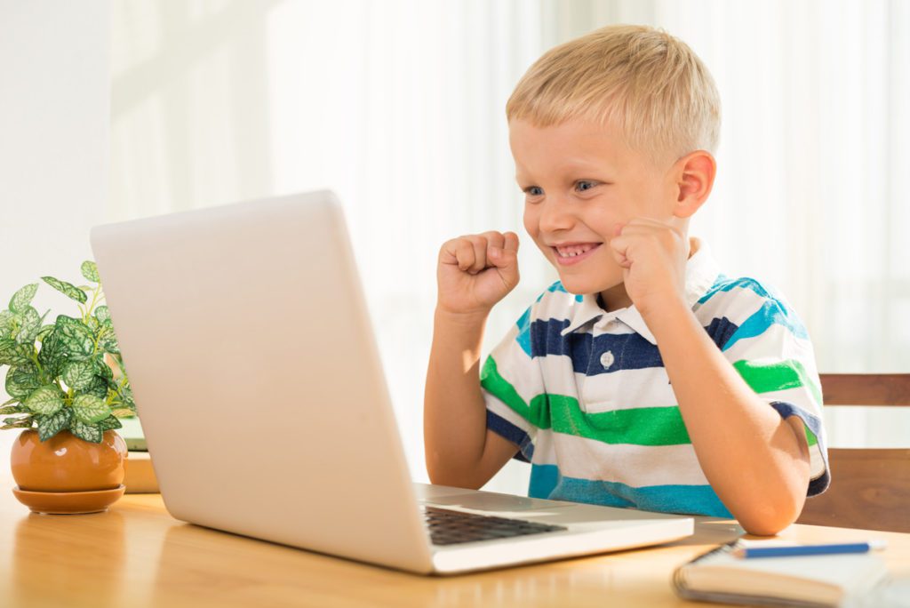 Elementary-age boy on a laptop at a kitchen table, smiling excitedly with his fists up.