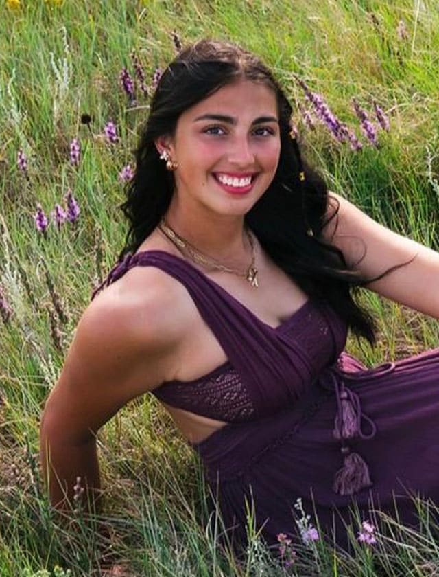 Smiling woman in a purple dress sitting in a grassy field with purple flowers surrounding her.