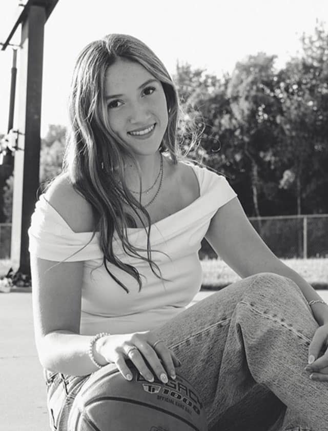 Smiling woman sits on a basketball court outdoors, with a ball near her knee and trees in the background.